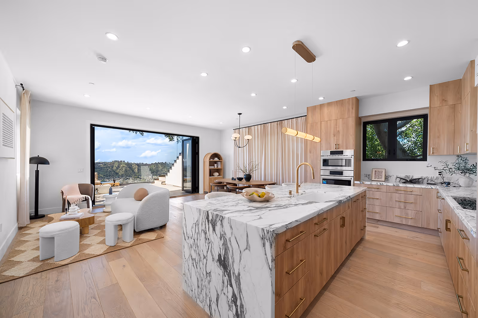 Modern open kitchen and living area with marble island, wood cabinetry, cozy seating, and large windows overlooking hillside view.