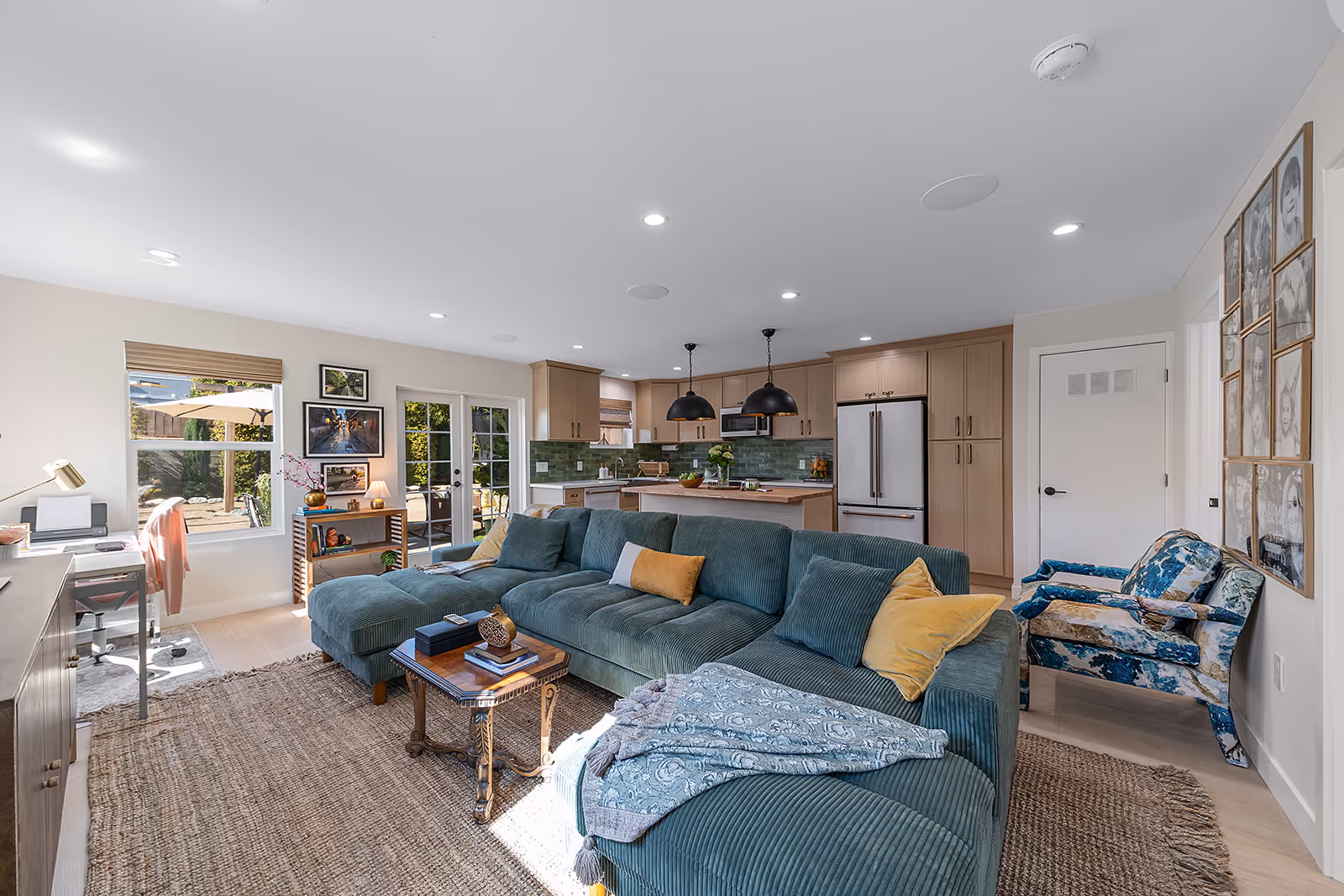 Bright open living room with teal sectional sofa, patterned armchair, wooden coffee table, and a kitchen with green tile backsplash.