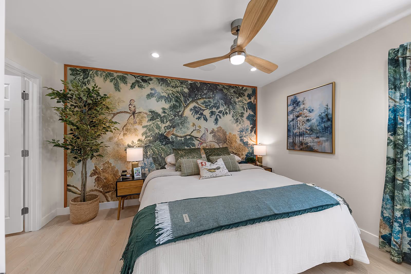 Modern bedroom with a large bed featuring green and beige pillows and throw, a tree mural on the back wall, bedside tables with lamps, a potted plant, and a ceiling fan with wooden blades.