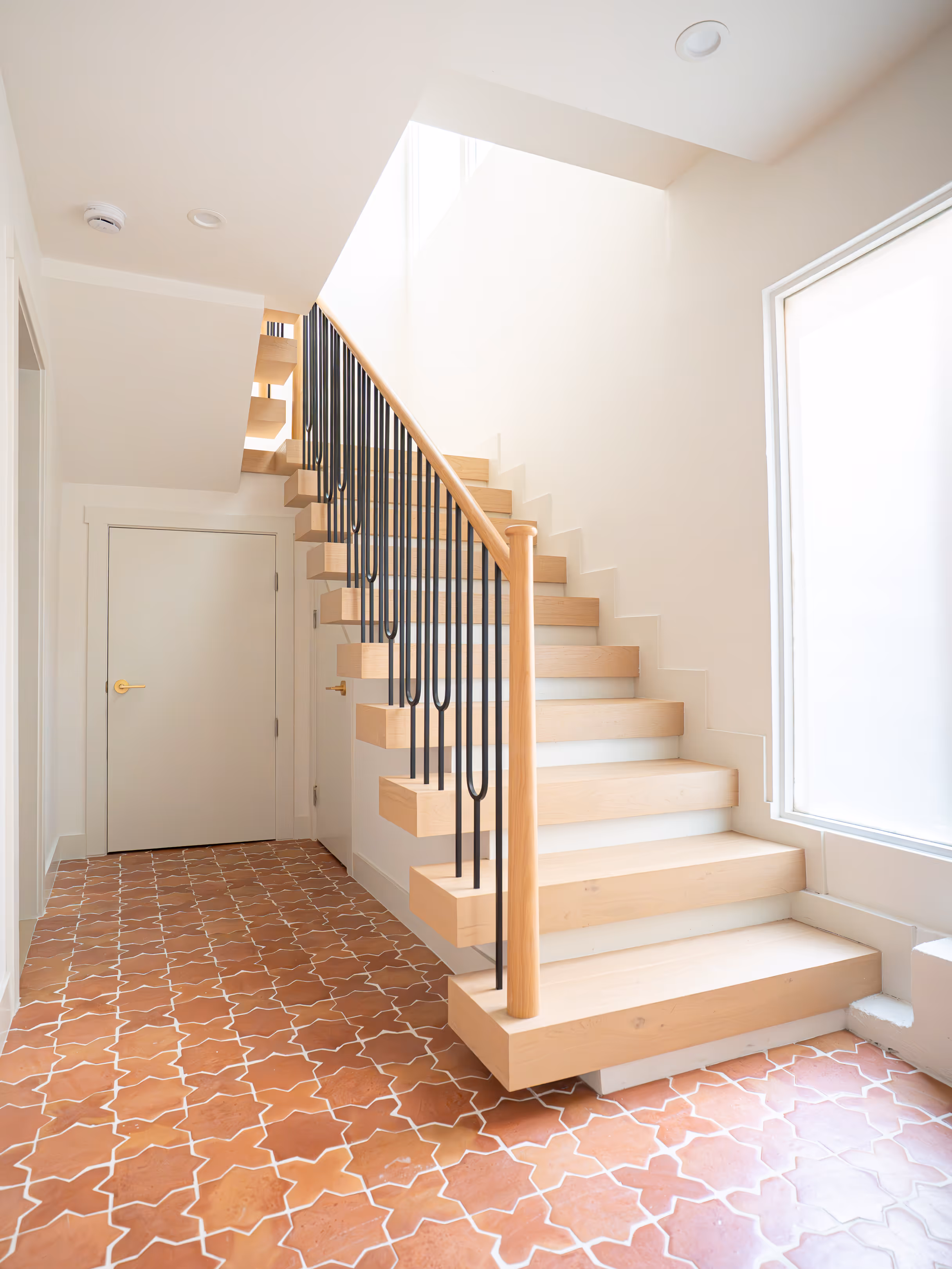 Bright modern hallway with light wood floating staircase, black metal railings, terracotta patterned tile floor, and white walls.
