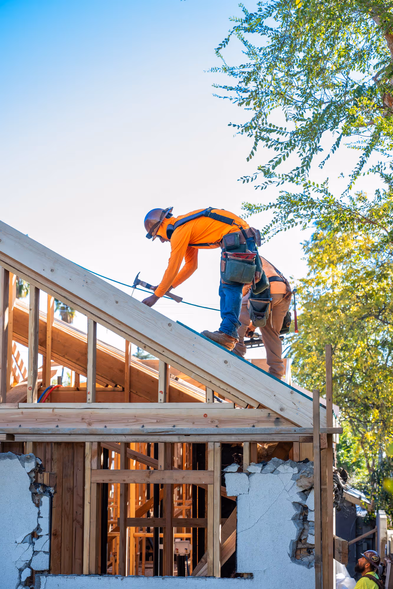 Construction workers wearing helmets and safety gear working on framing and roofing of a wooden house under clear sky.