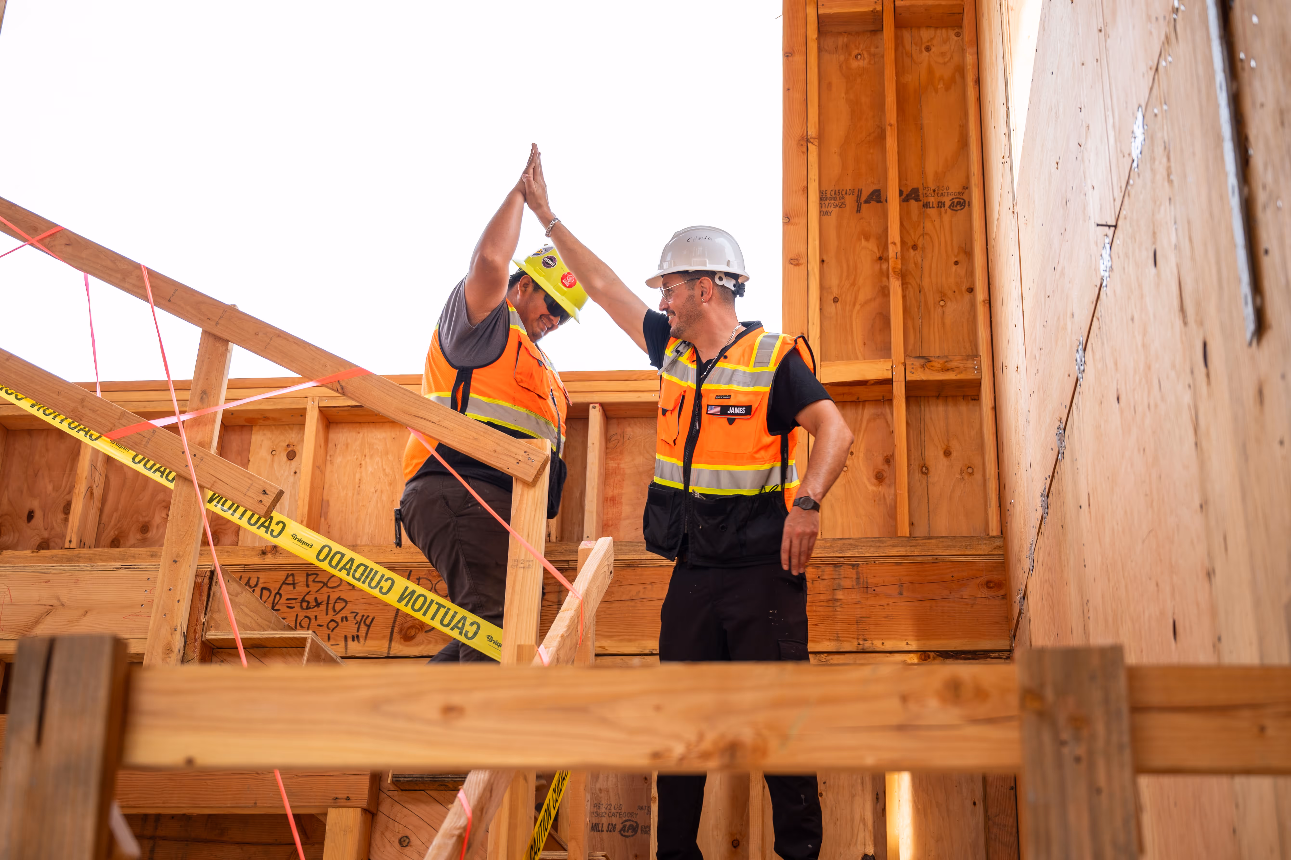Two construction workers in safety vests and helmets high-five on a wooden building frame.