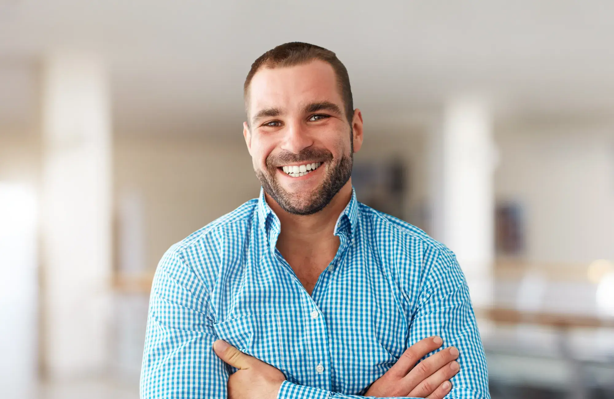 Smiling man with a beard wearing a blue checkered shirt, standing with arms crossed indoors.