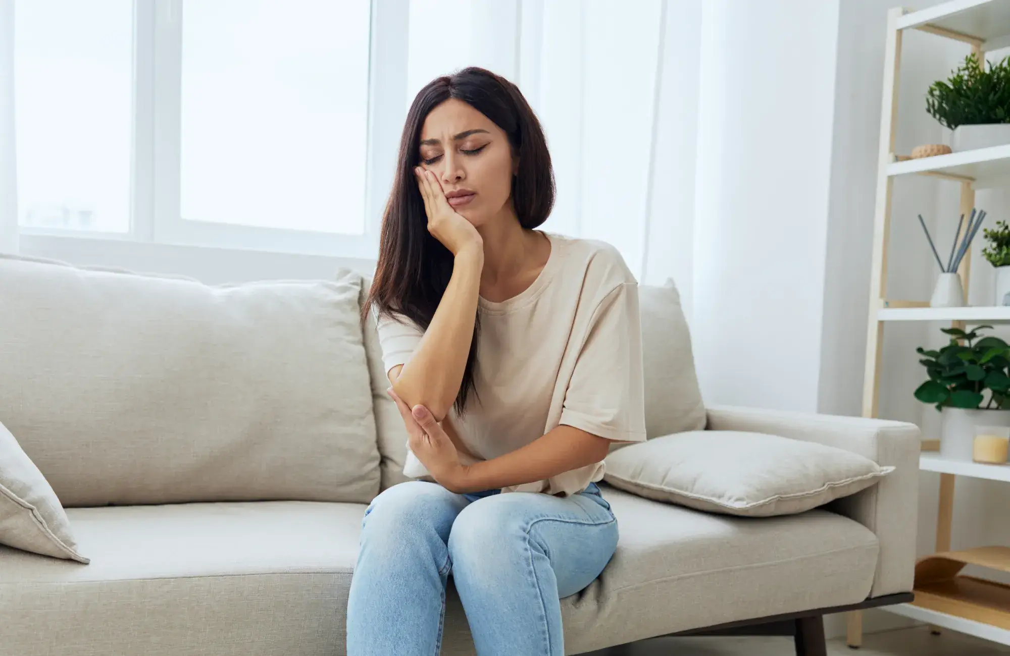Woman sitting on a beige sofa holding her cheek and grimacing in pain, indicating a toothache.