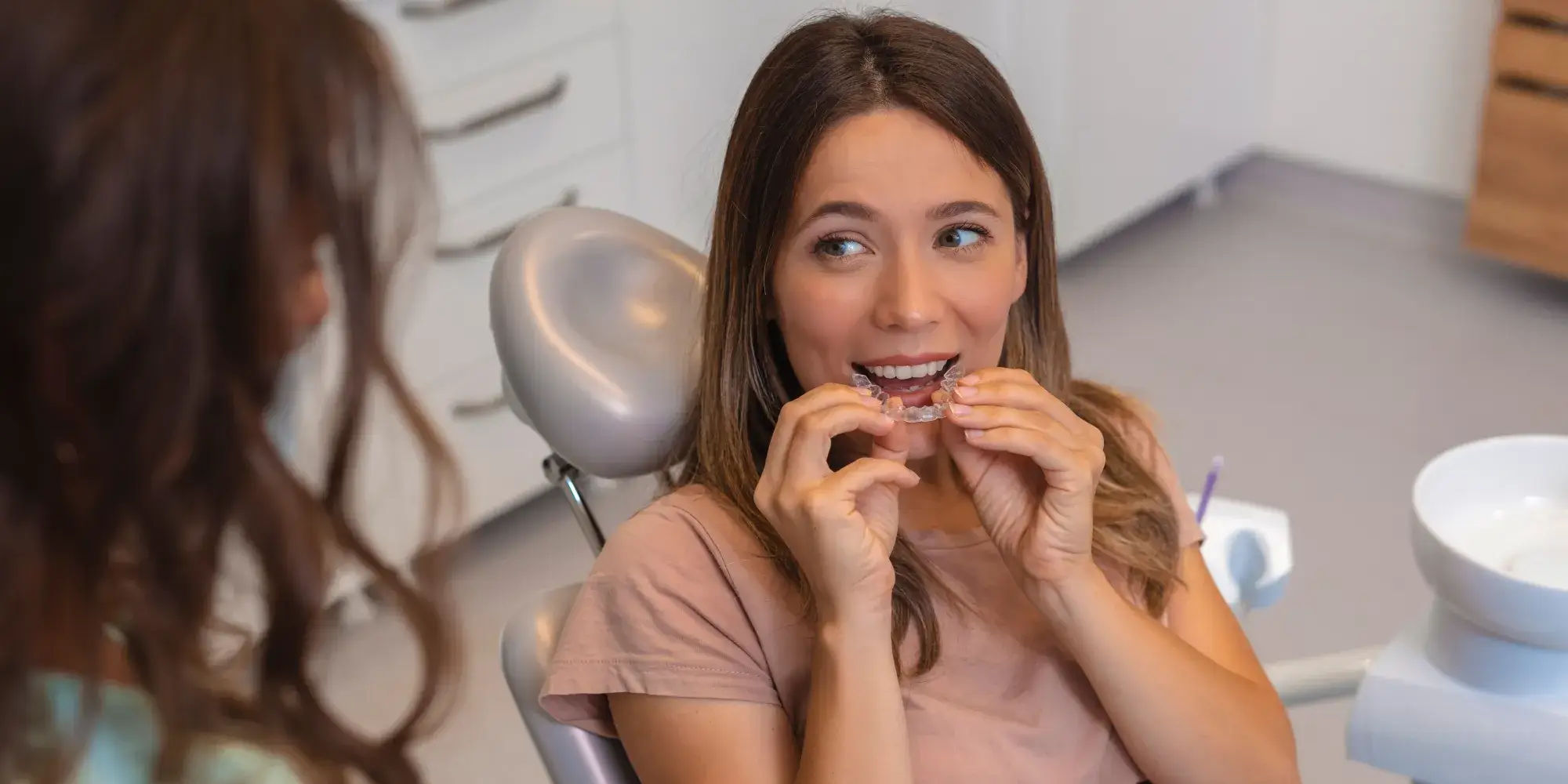 Woman sitting in a dental chair holding a clear orthodontic retainer, with a dentist nearby.