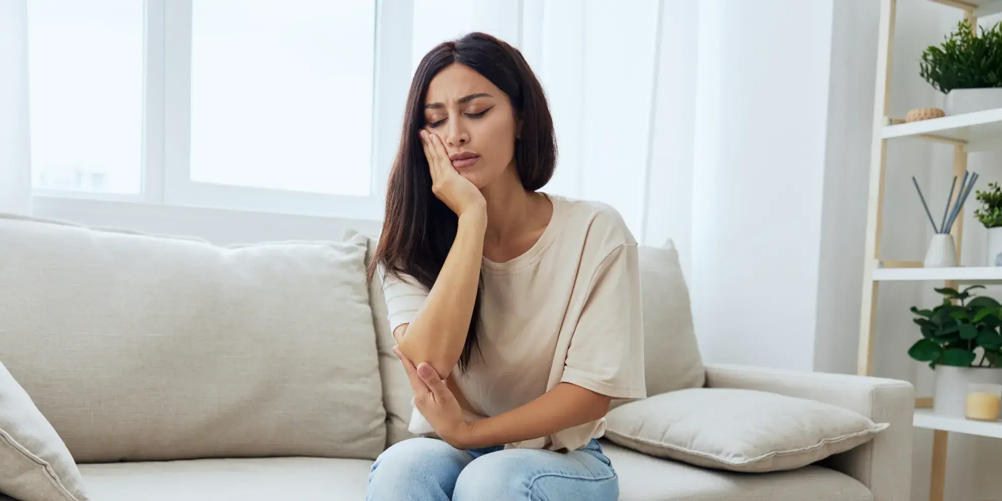 A woman sits on a couch, holding her cheek with a pained expression.