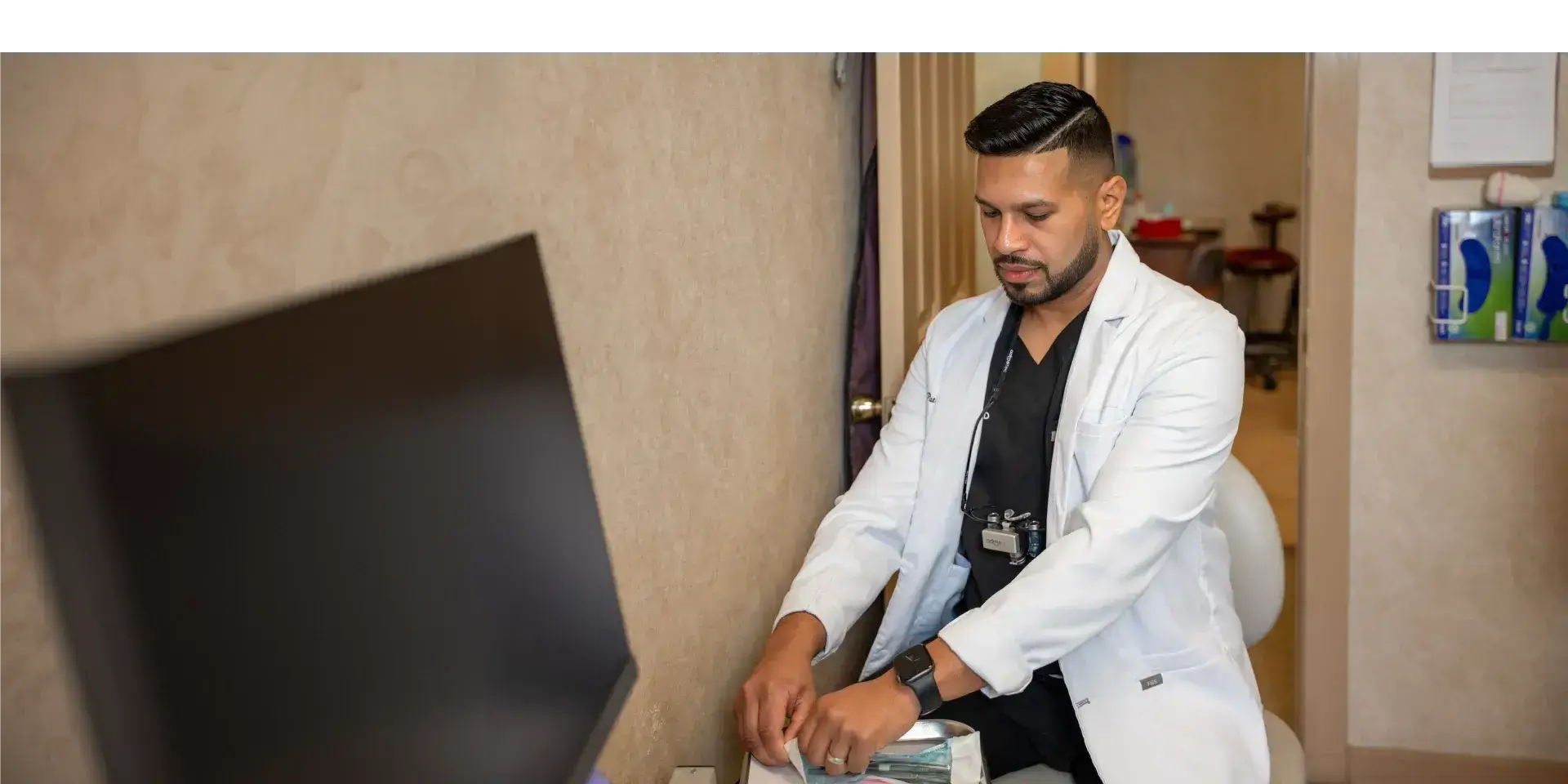 Male healthcare professional in white coat preparing medical supplies while seated in a clinical room.