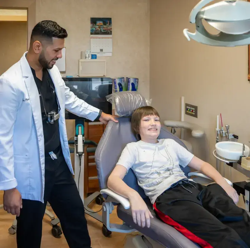 Smiling boy reclines in dental chair while dentist in white coat stands nearby.
