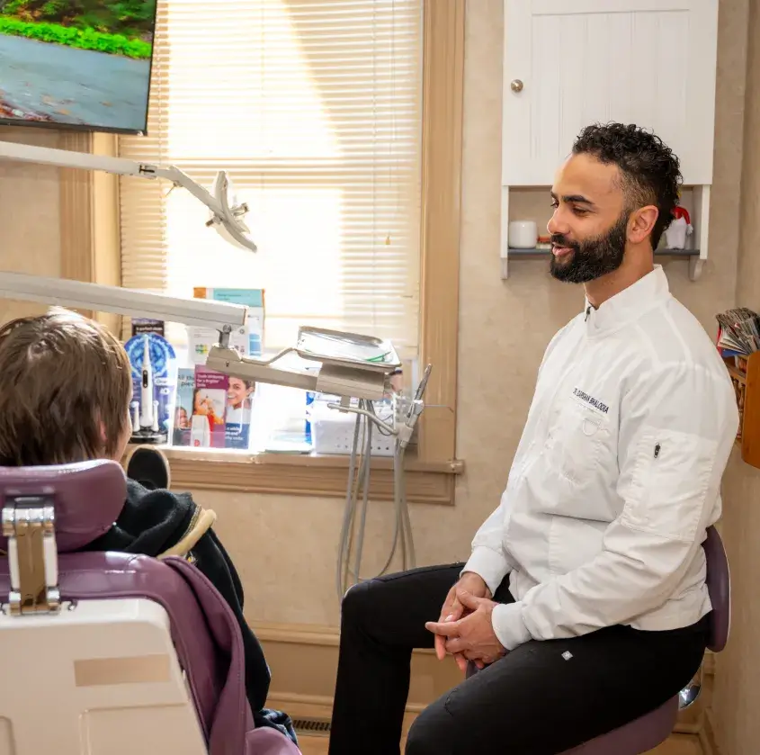 Dentist in white coat sitting and talking to patient in dental chair in bright office.