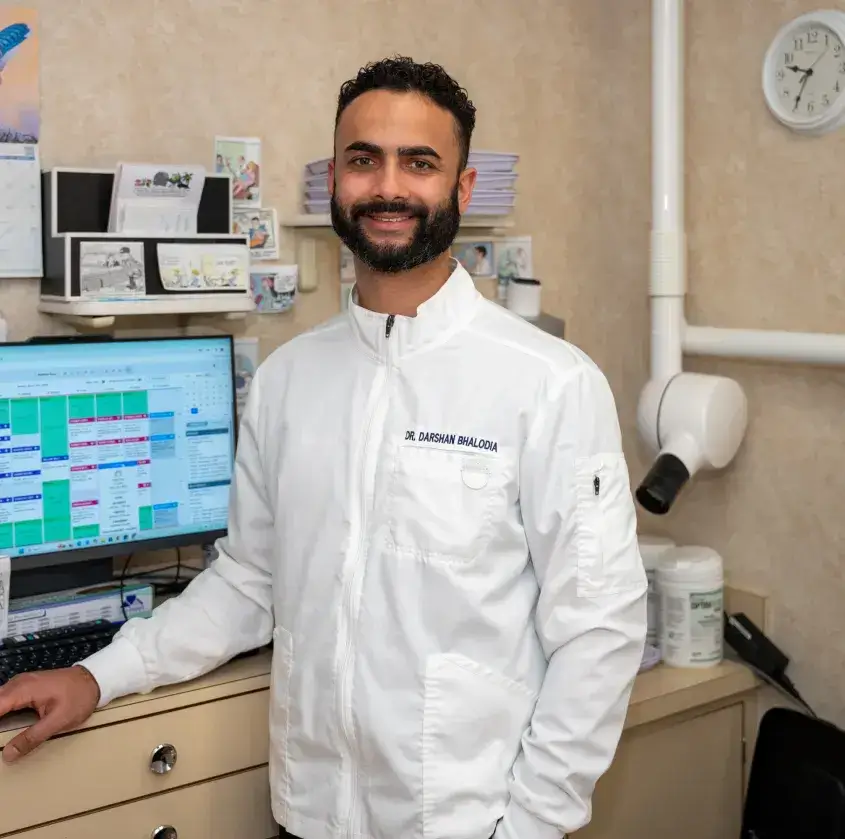 Smiling male dentist Dr. Darshan Bhalodia in white coat standing in office with computer and dental equipment.