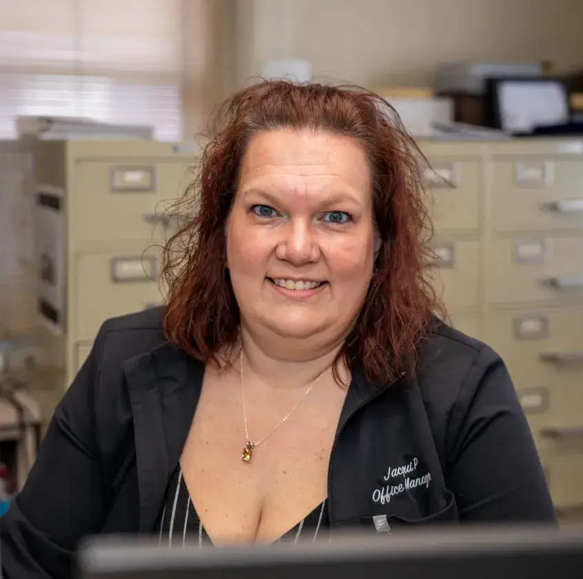 Smiling woman with red hair wearing a jacket labeled Office Manager, seated in an office with filing cabinets.