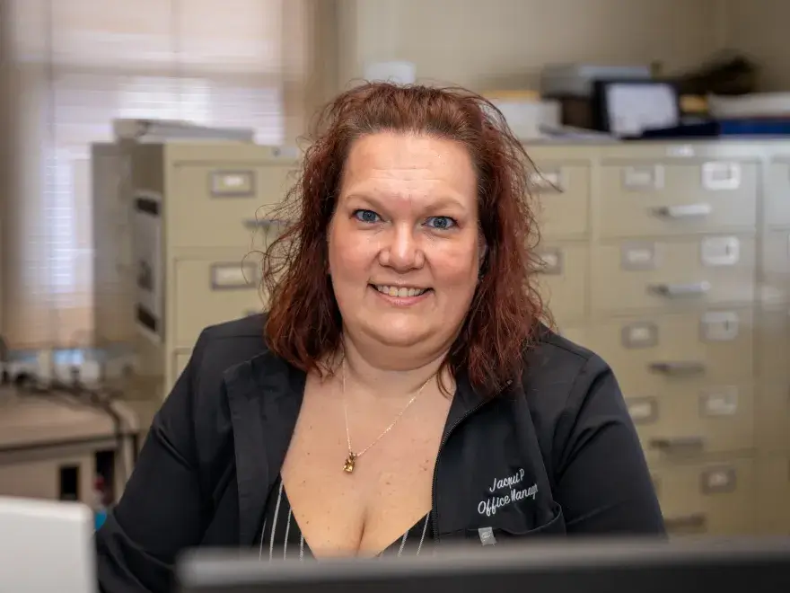 Smiling woman with auburn hair sitting at a desk in front of filing cabinets in an office.