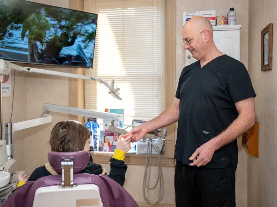 Dentist handing a toothbrush to a child seated in a dental chair in a clinic room.