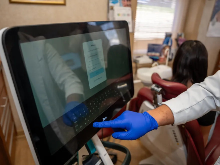 Gloved hand of a medical professional using a touchscreen device near a patient in a dental chair.