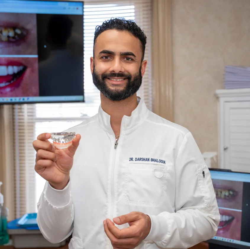 Smiling male dentist in white coat holding a dental mold with images of teeth on monitors behind.