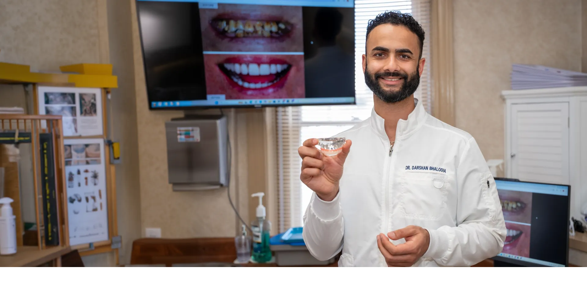Smiling dentist in white coat holding clear dental mold in office with teeth images on screens.
