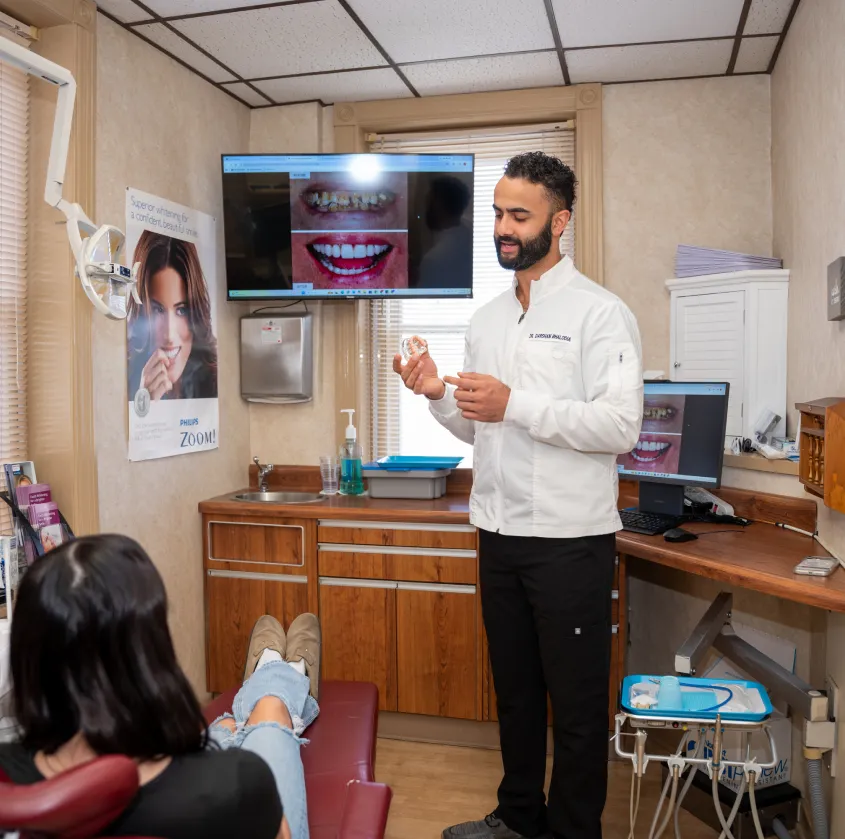 Dentist showing dental model to patient in clinic with before-and-after teeth images on screen.