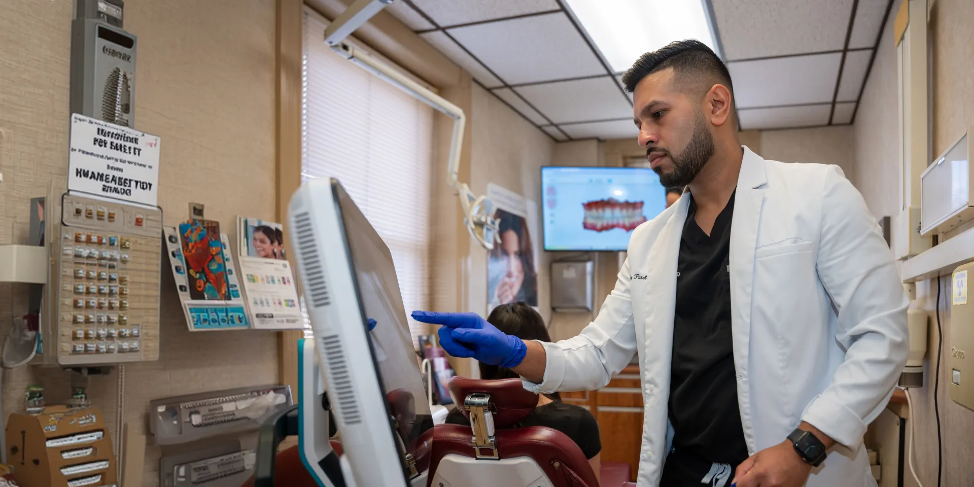Dentist wearing gloves pointing at a screen while a patient sits in a dental chair.