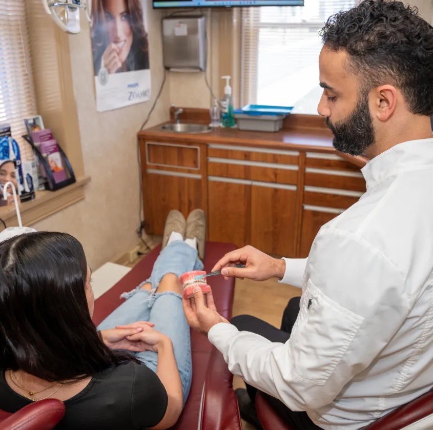 Dentist demonstrating proper brushing on a dental model to a seated patient in a clinic.