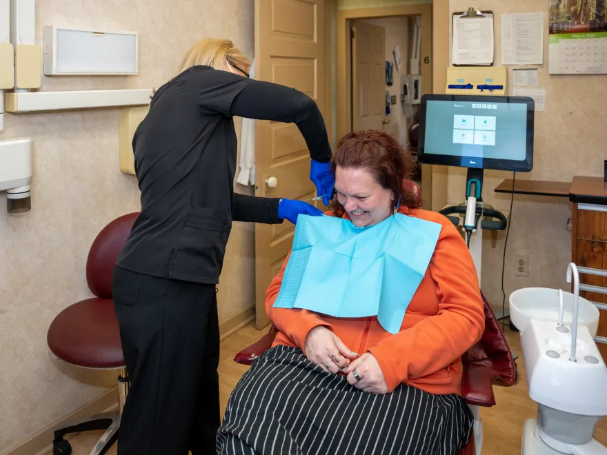 Dental hygienist wearing blue gloves cleaning woman’s teeth in an exam room with dental equipment.
