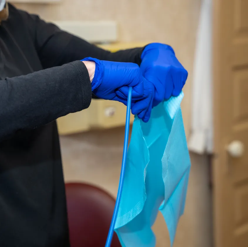 Person wearing blue gloves holding a blue dental bib and a dental suction tool.