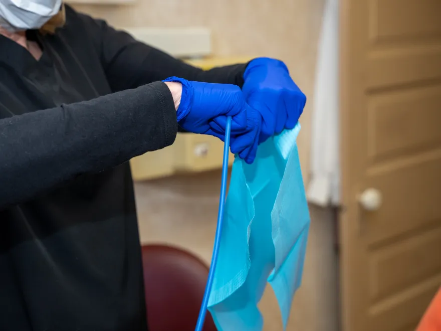 Person wearing blue gloves and black sleeves holding a blue dental bib and dental suction tube.