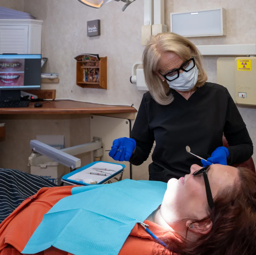 Dentist wearing gloves and mask examining a patient lying in dental chair with tools nearby.
