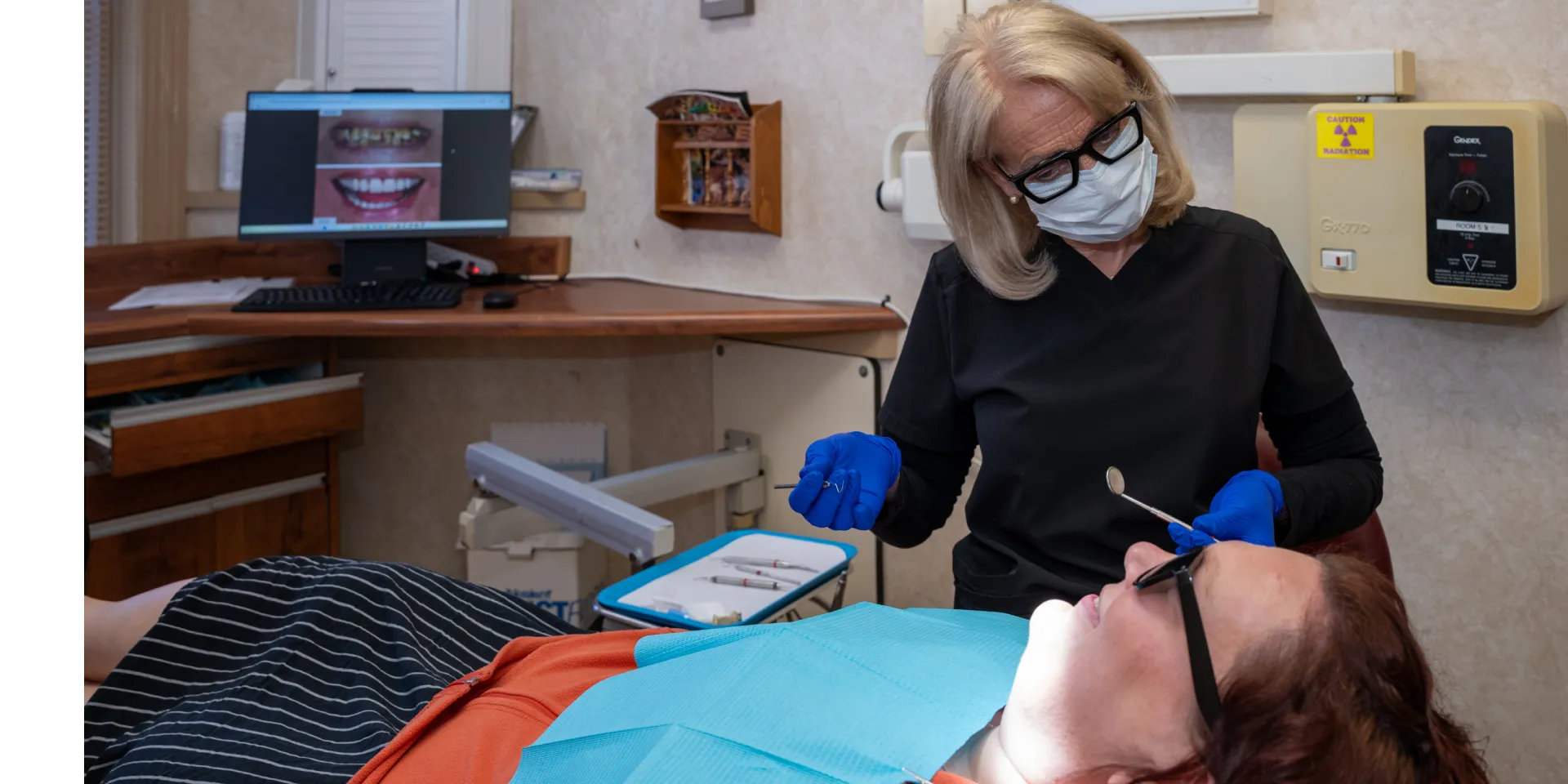 Dentist wearing mask and gloves examining a patient lying in a dental chair with oral tools.