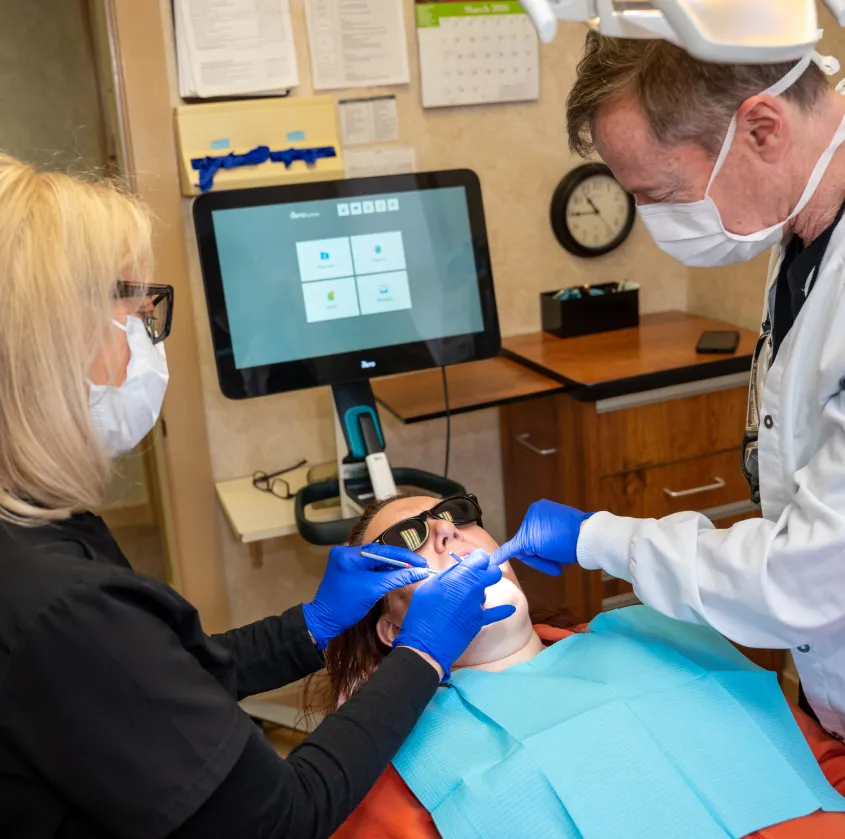 Dentist and assistant wearing masks and gloves treating patient reclining in chair with dental tools.