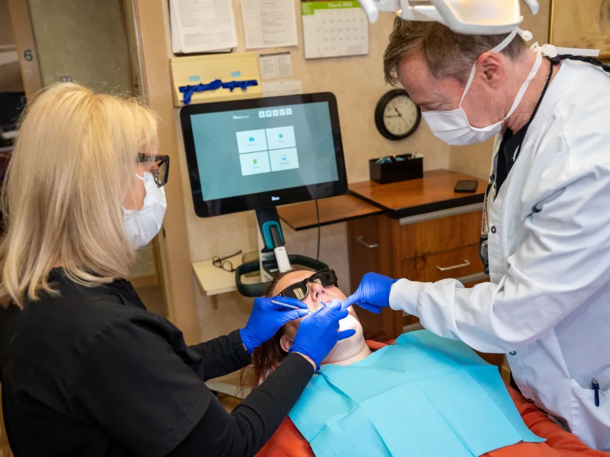 Dentist and assistant wearing masks and gloves treating a patient reclining in a dental chair.