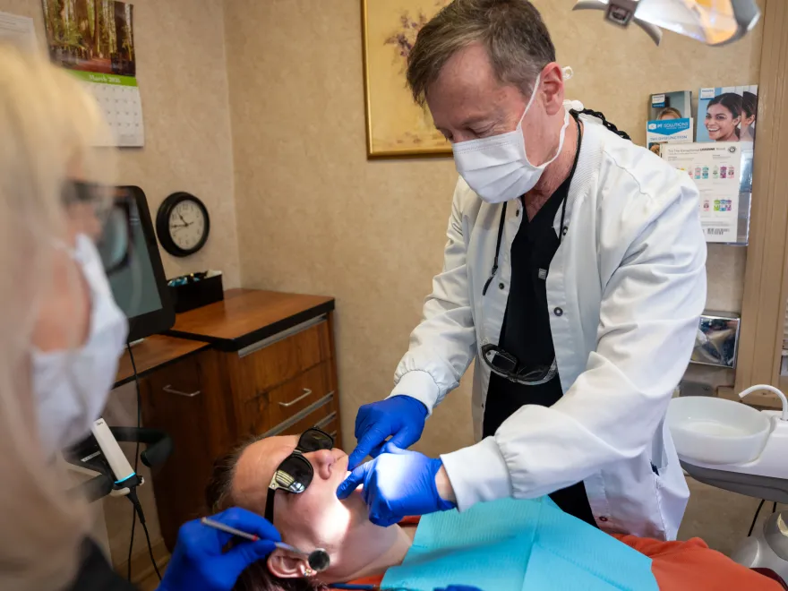 Dentist wearing mask and gloves treating patient wearing sunglasses in dental clinic.