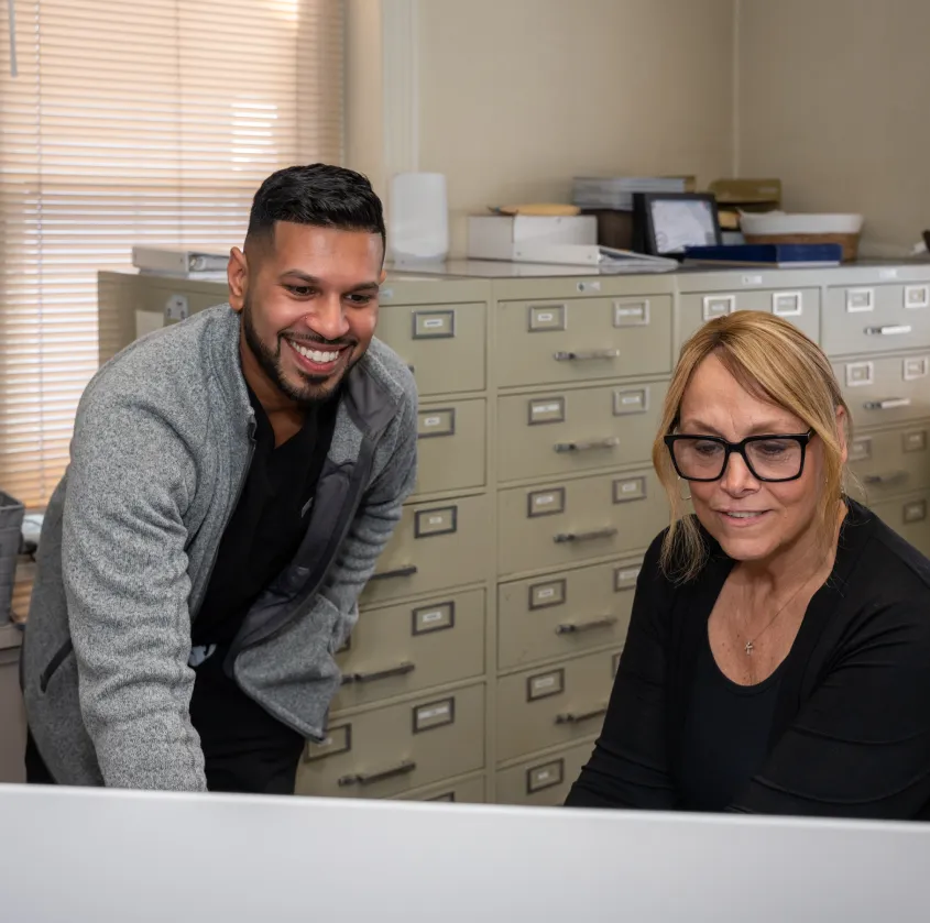 Two coworkers looking at a computer screen in an office with filing cabinets in the background.