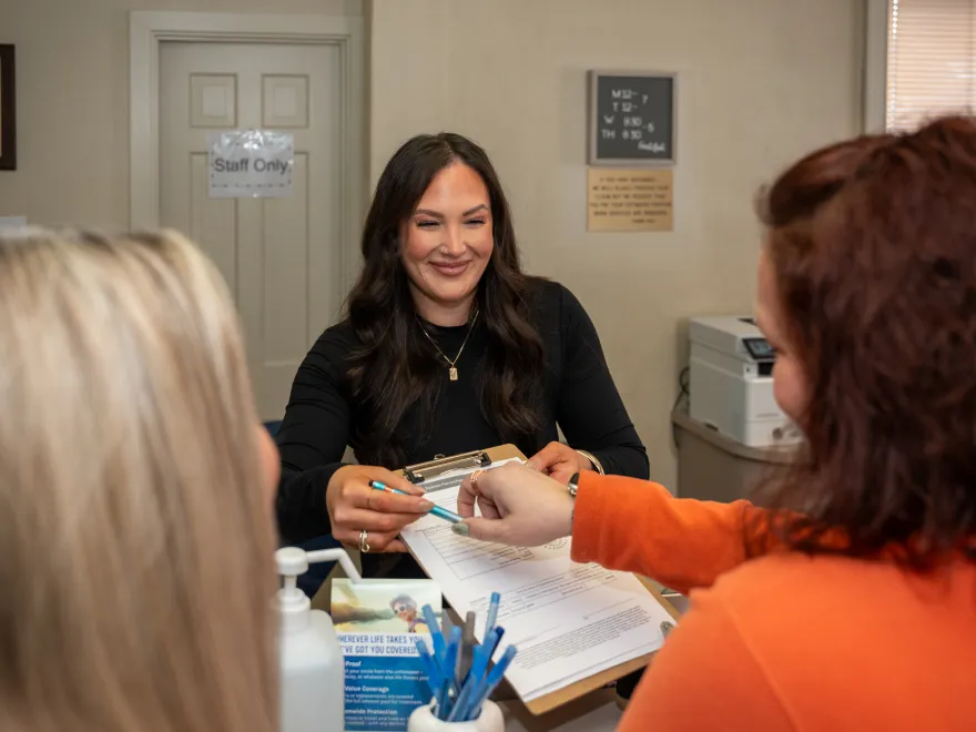 Smiling woman behind counter hands clipboard to another woman holding a pen in an office setting.