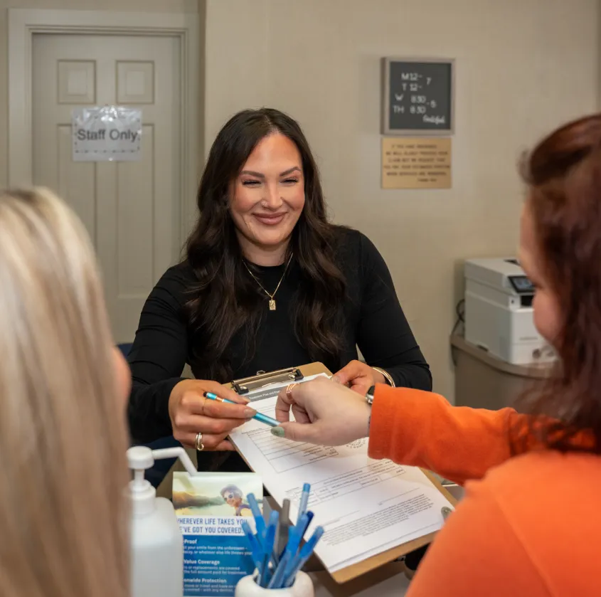 Smiling woman with clipboard assisting two clients in an office setting with form and pens.