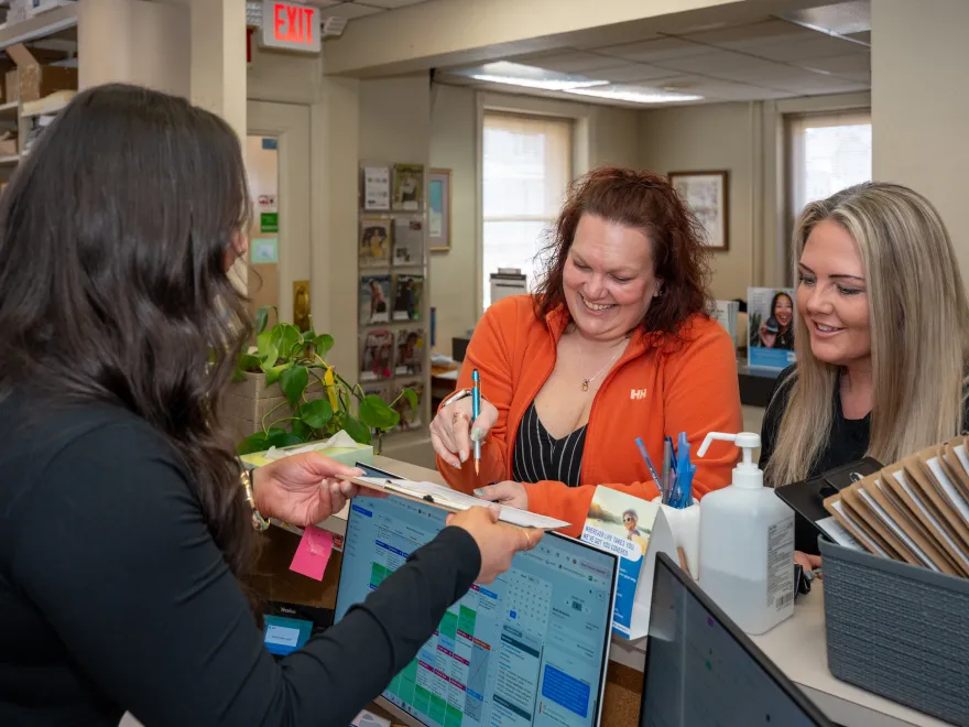 Two women smiling and signing papers across a desk from a woman with long dark hair working on a laptop.