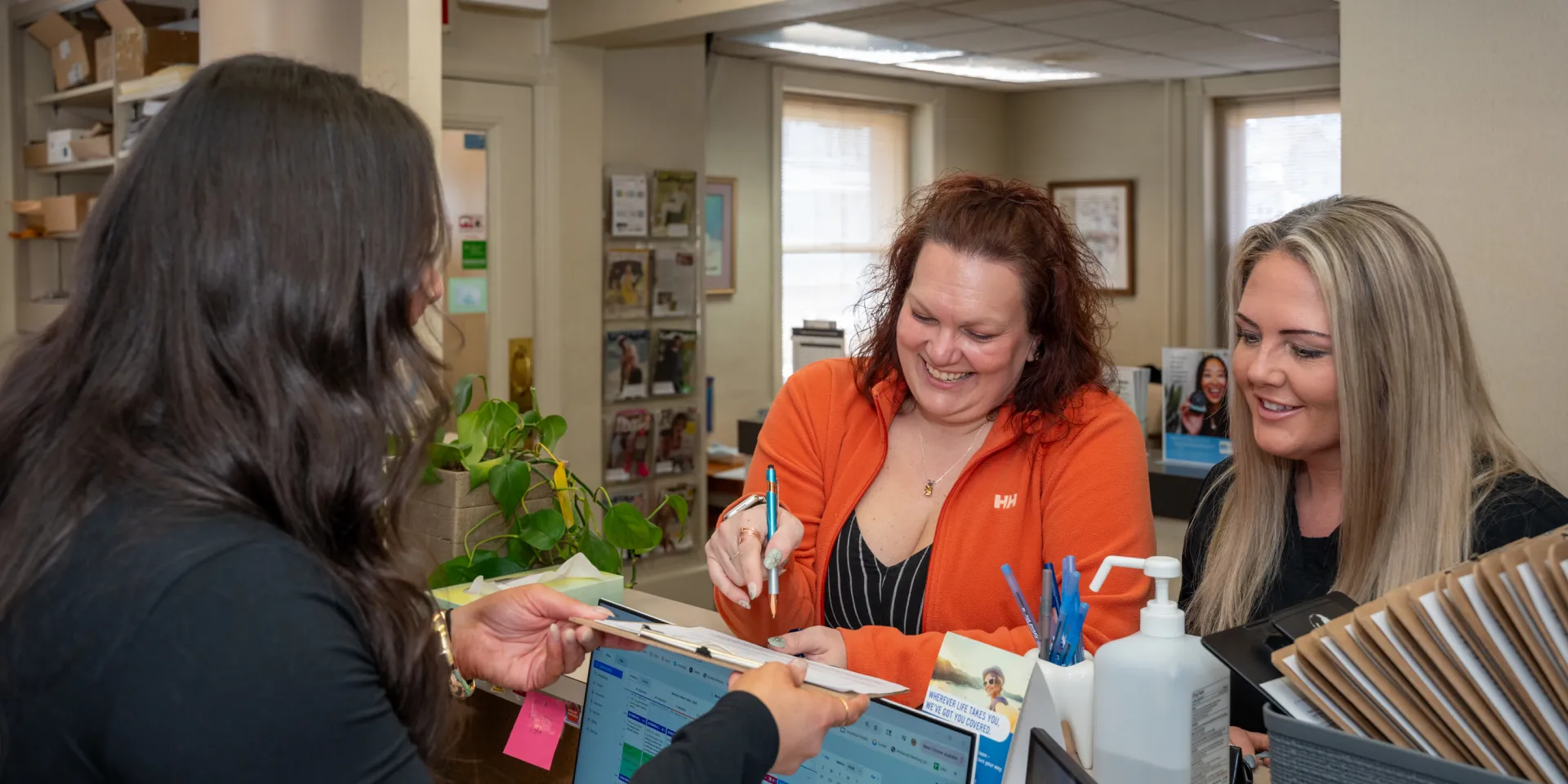 Two women at a desk smiling interact with a woman holding a clipboard in an office setting.
