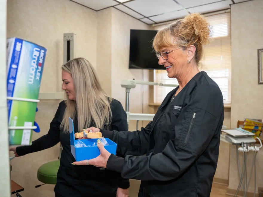 Two women in black scrubs smiling and handling dental molds in a dental office.
