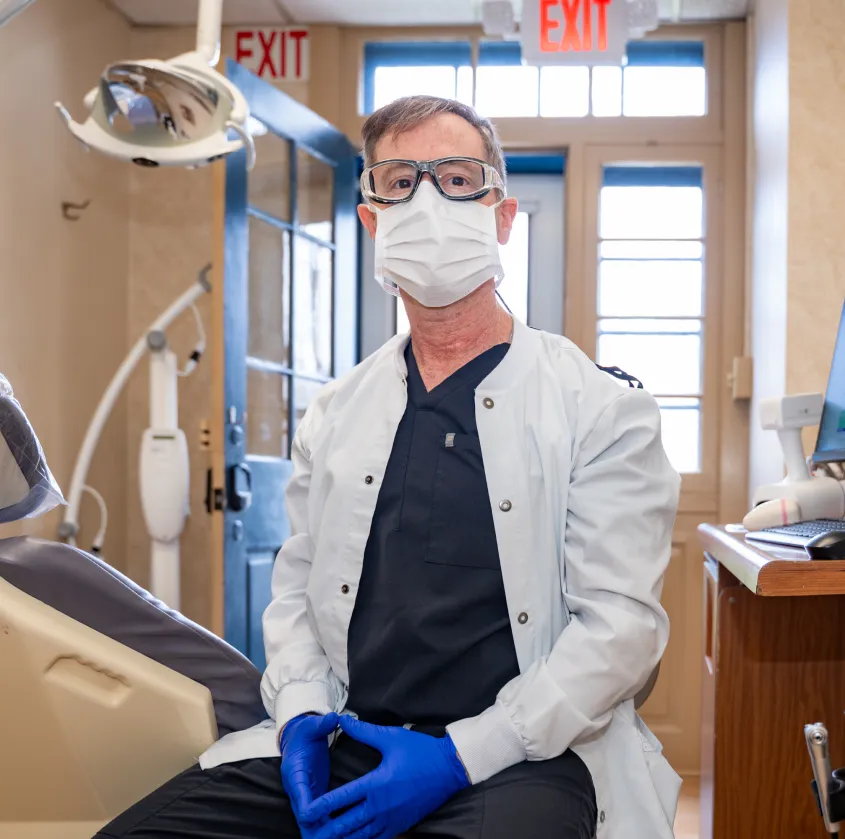Dentist wearing mask, goggles, gloves, and lab coat sitting in dental clinic with equipment visible.