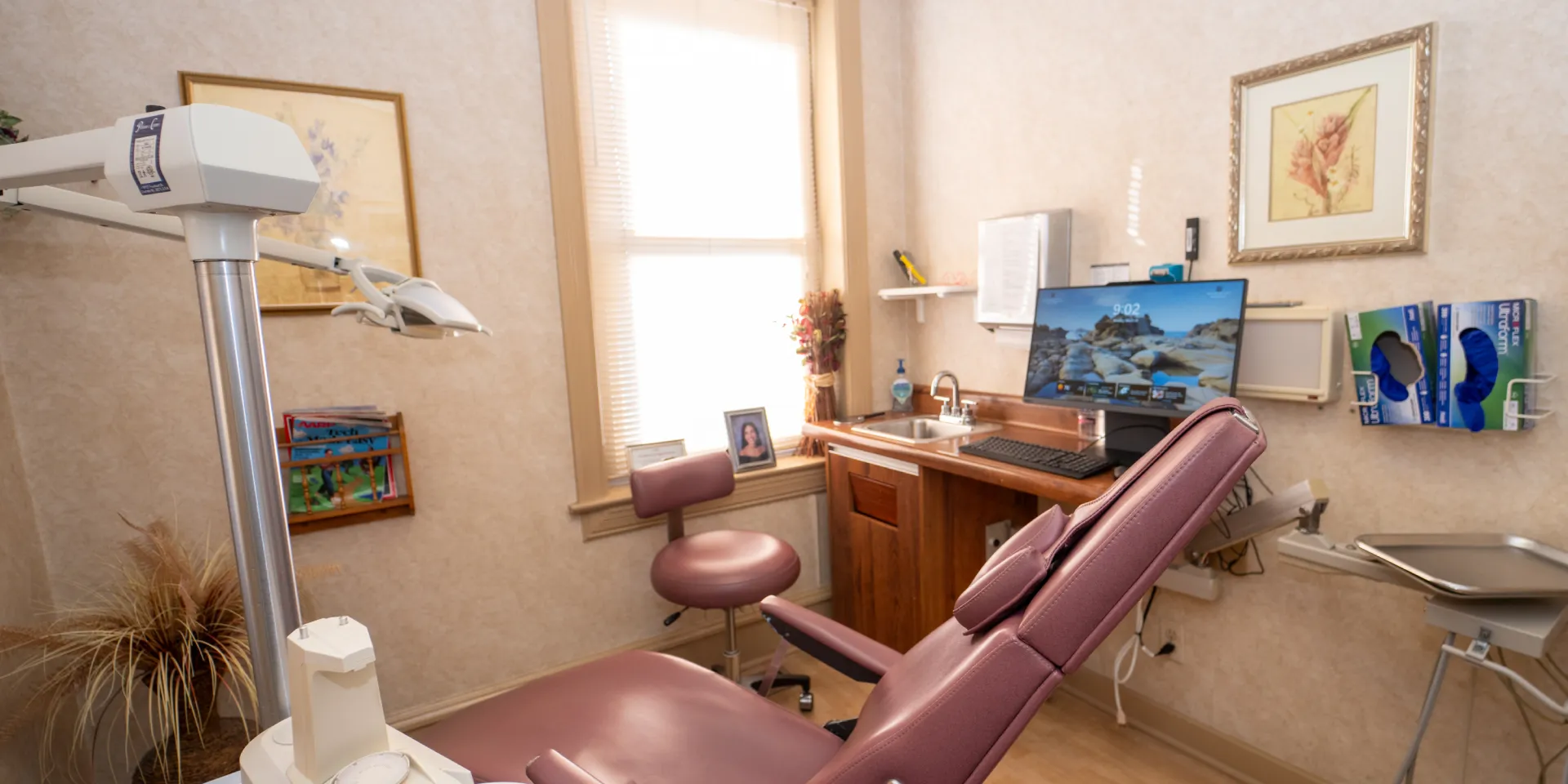 Dental examination room with maroon patient chair, overhead light, sink, and computer on counter.