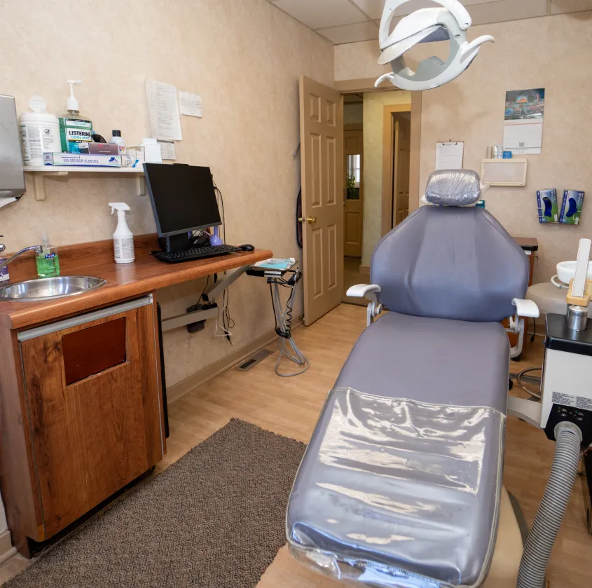Empty dental exam room with gray dental chair, overhead light, computer, sink, and medical supplies.