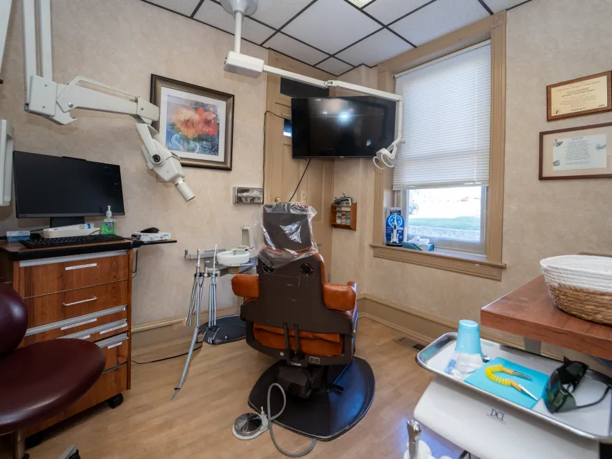 Dental office with a chair, overhead light, monitor, and dental tools near a window with certificates on the wall.