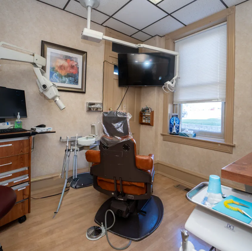 Dental clinic room with patient chair, overhead light, TV, and dental instruments near a window.