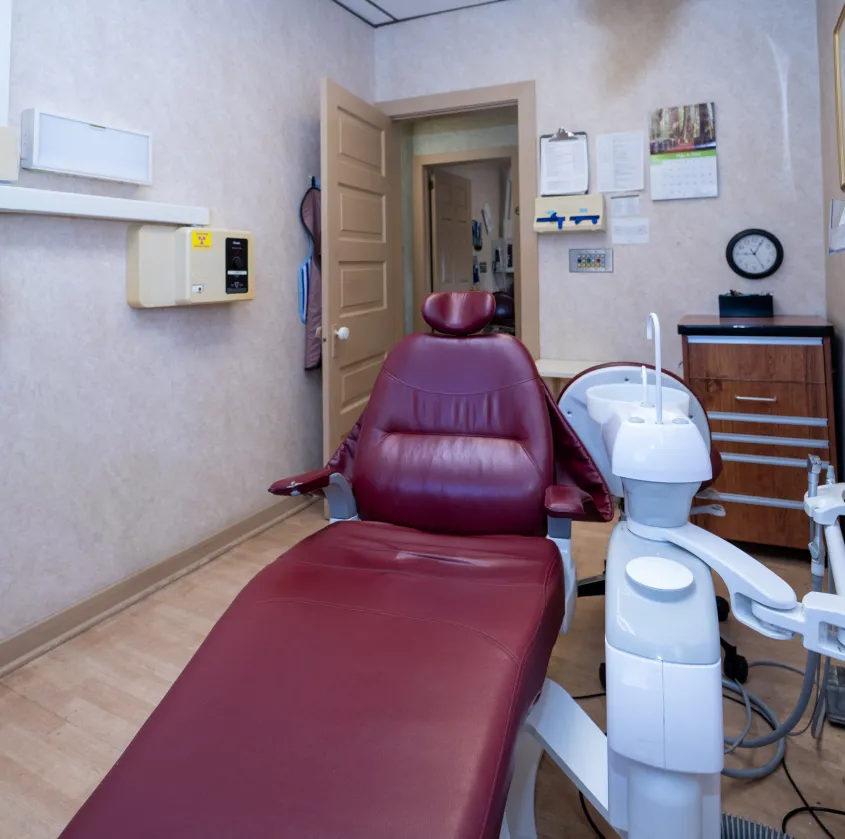 Empty dental chair in a clinic room with equipment, wooden cabinet, and open door in the background.