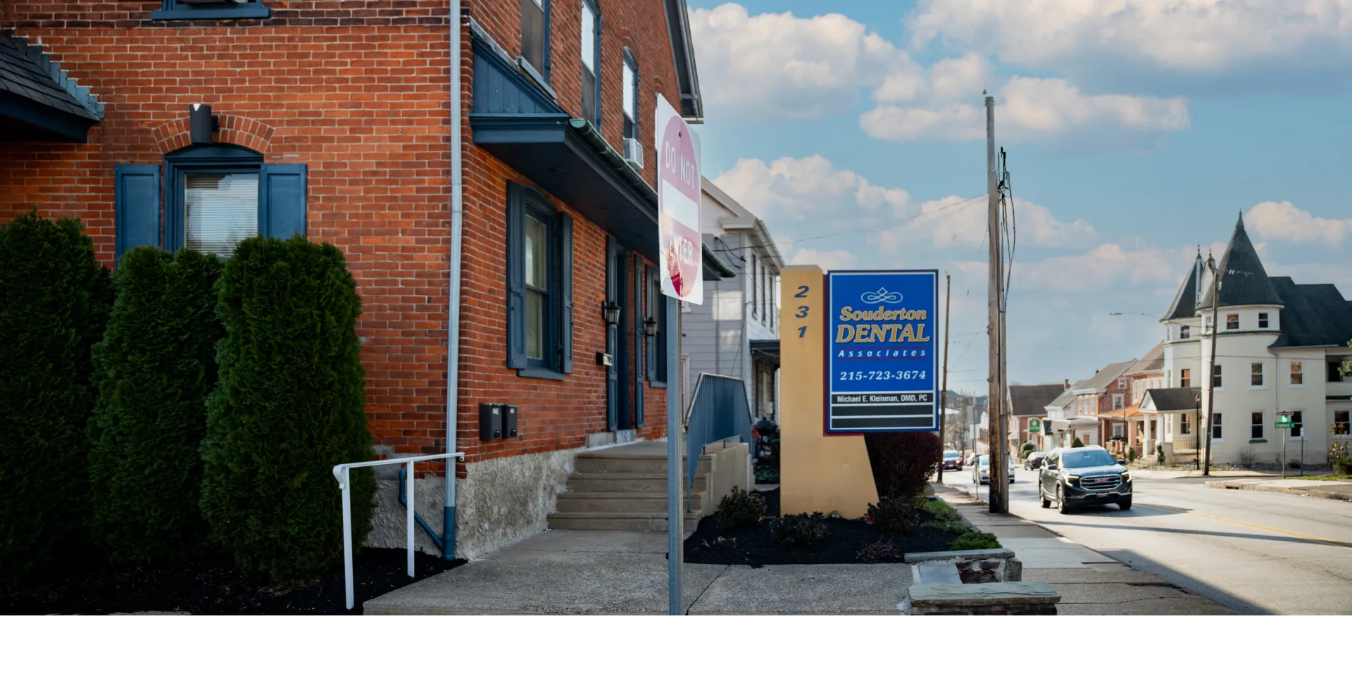 Street view of brick building with blue windows and a Souderton Dental Associates sign by the sidewalk.