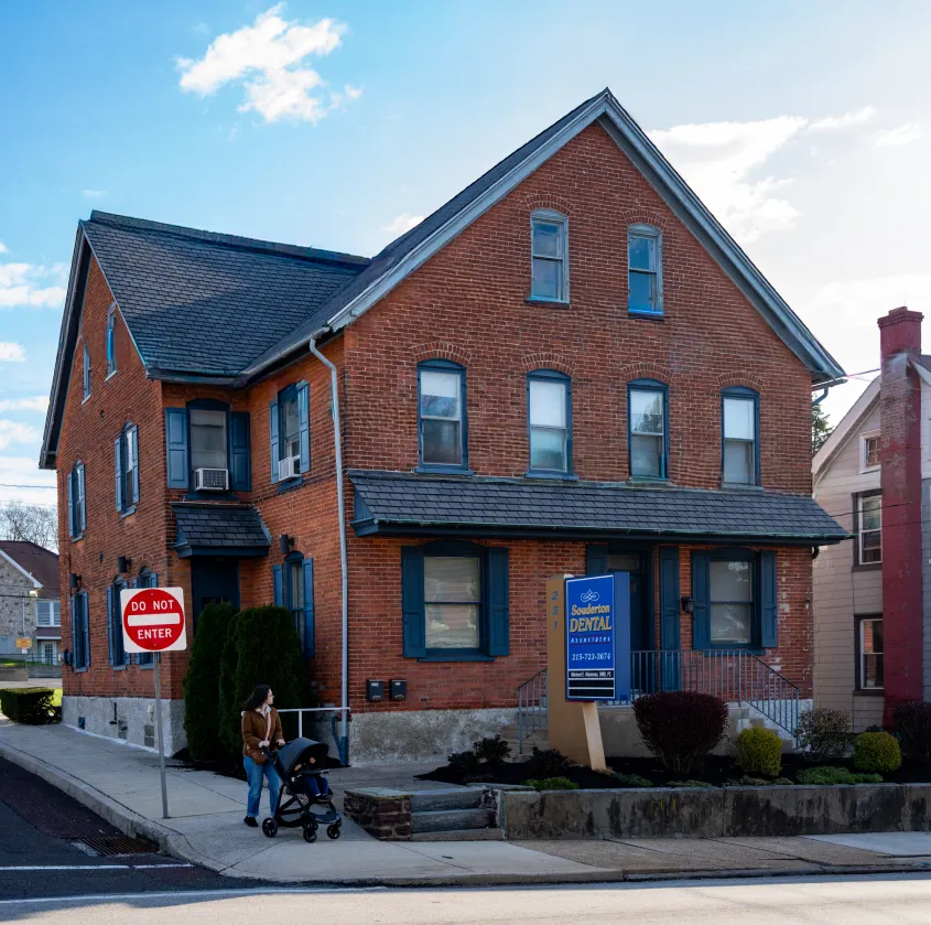 Red brick building with blue shutters and a dental office sign, woman walking with stroller nearby.