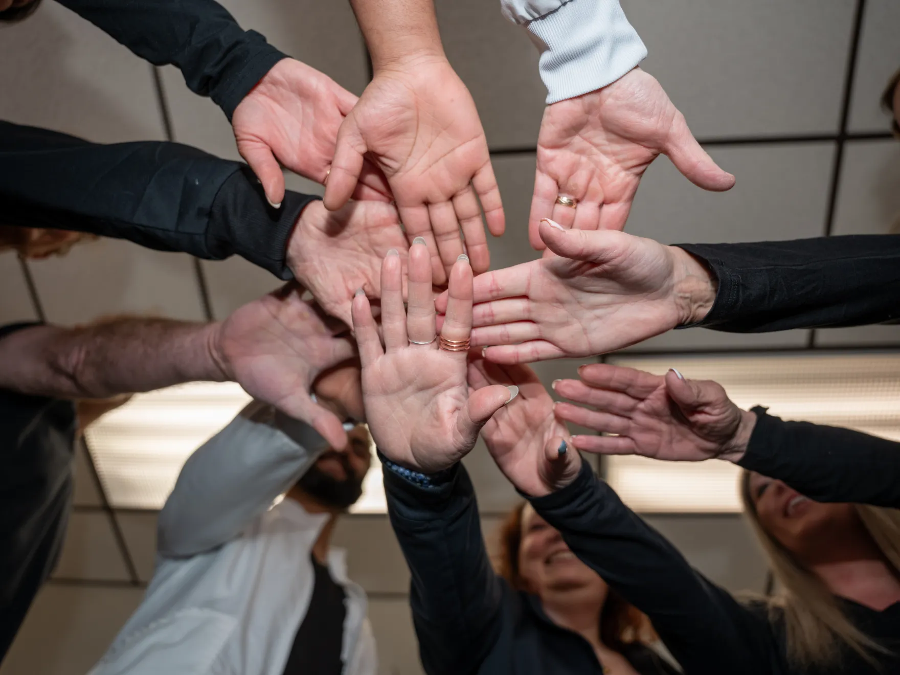 Group of people stacking hands in a team gesture, viewed from below with smiling faces around.
