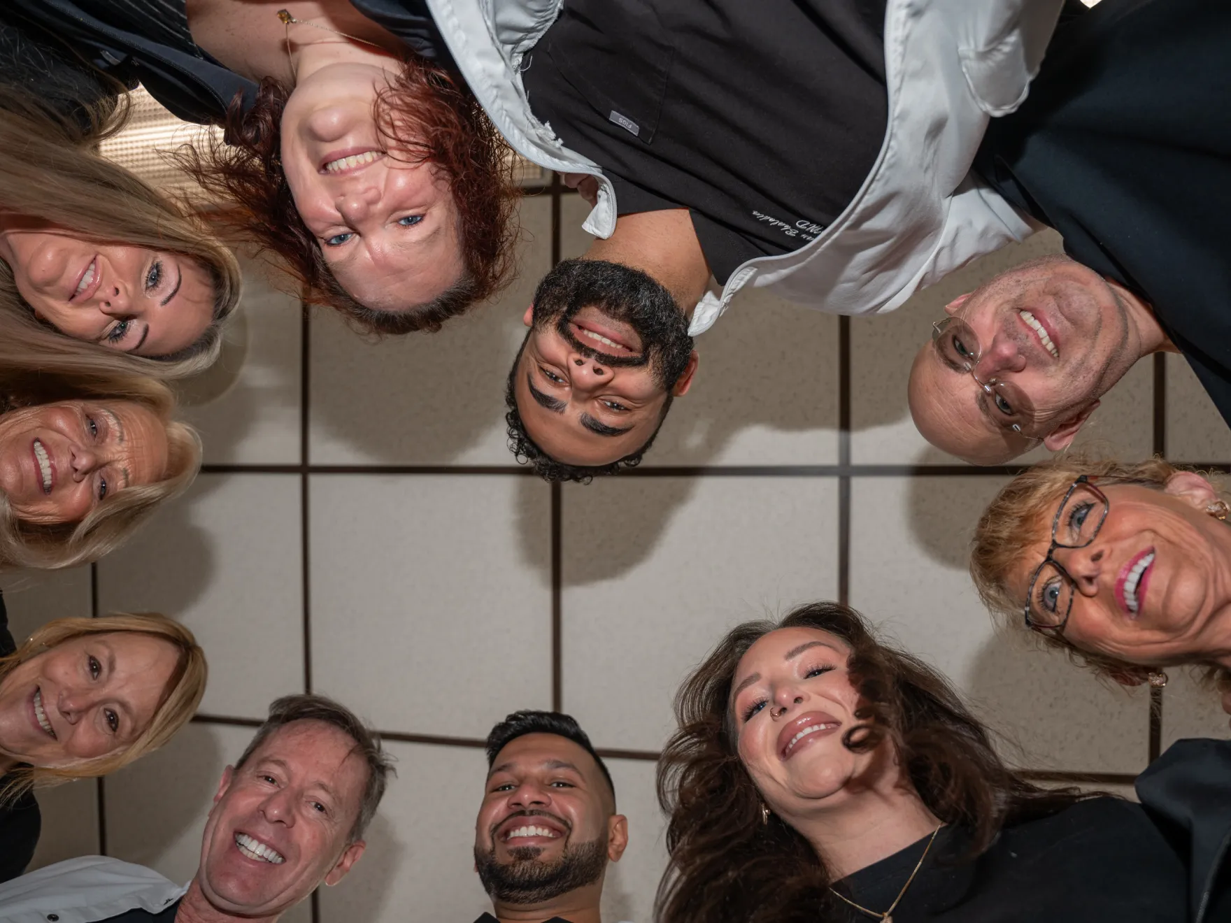 Group of ten people standing in a circle smiling and looking down at the camera indoors.