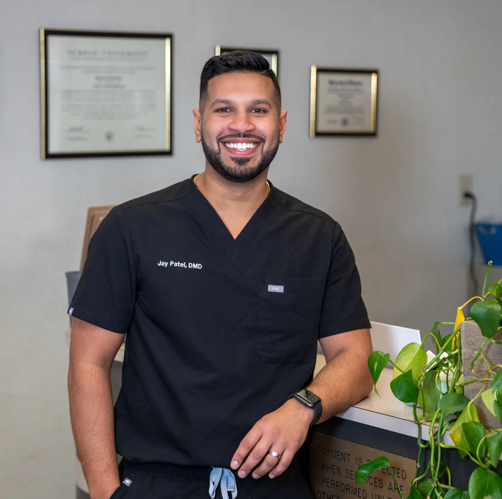 Smiling man in black scrubs with name Jay Patel, DMD, standing in office with framed certificates behind him.