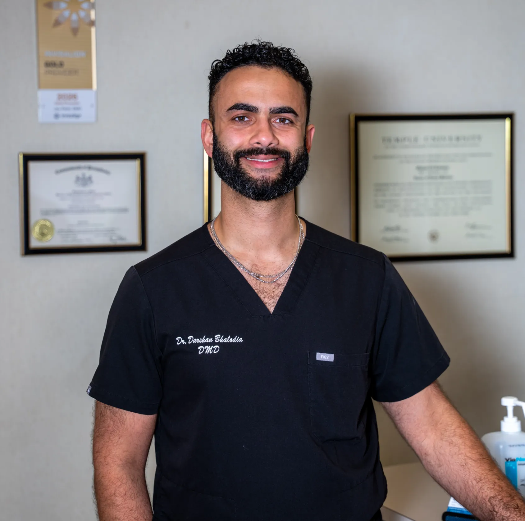 Smiling man with beard wearing black scrubs standing in front of framed certificates on wall.