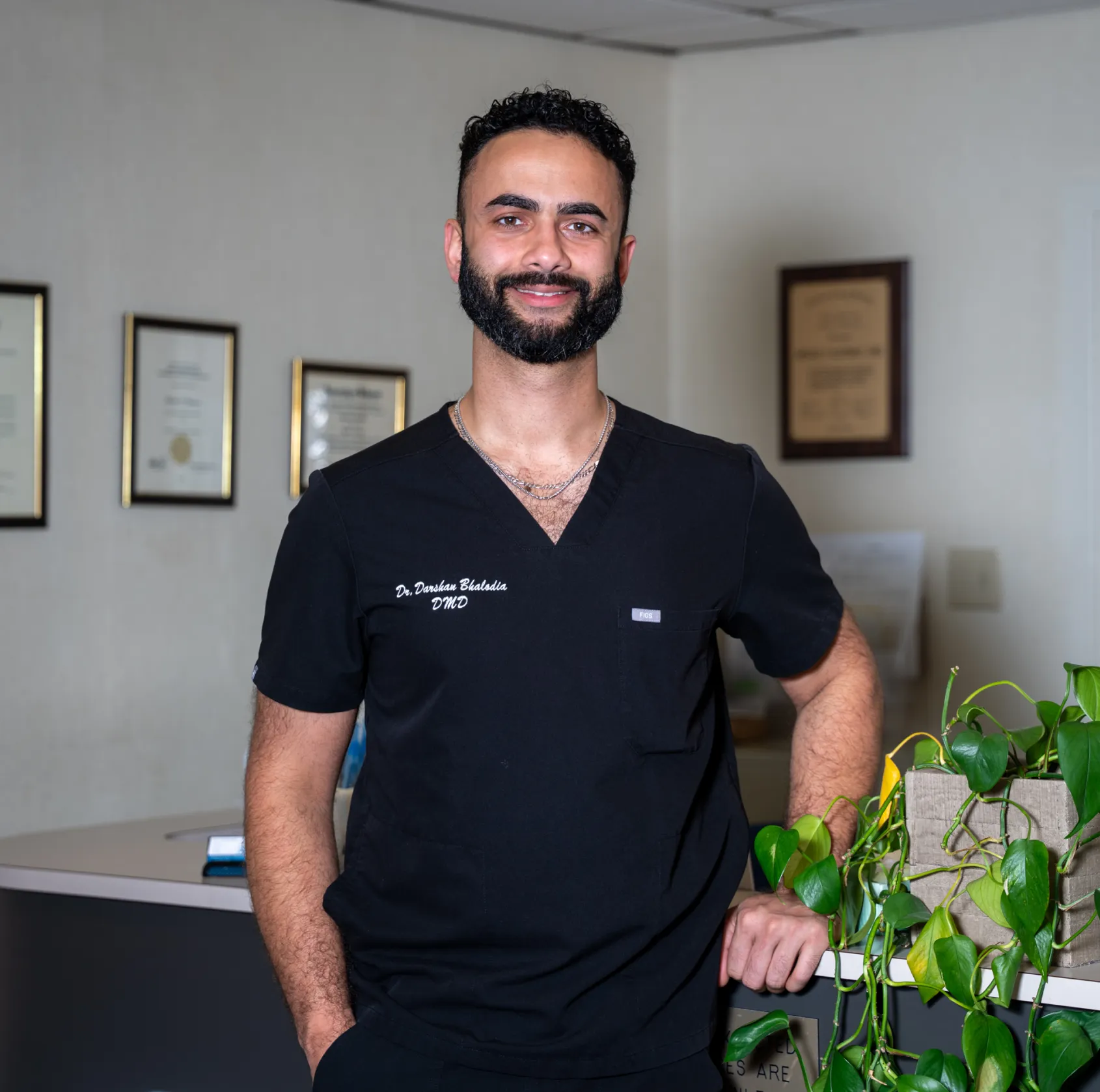 Smiling male doctor in black scrubs leans on counter with green plant and framed certificates behind.